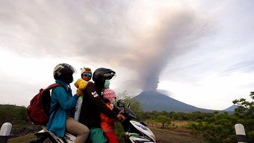 A family on a motorcycle passes-by the Mount Agung volcano erupting in the background.