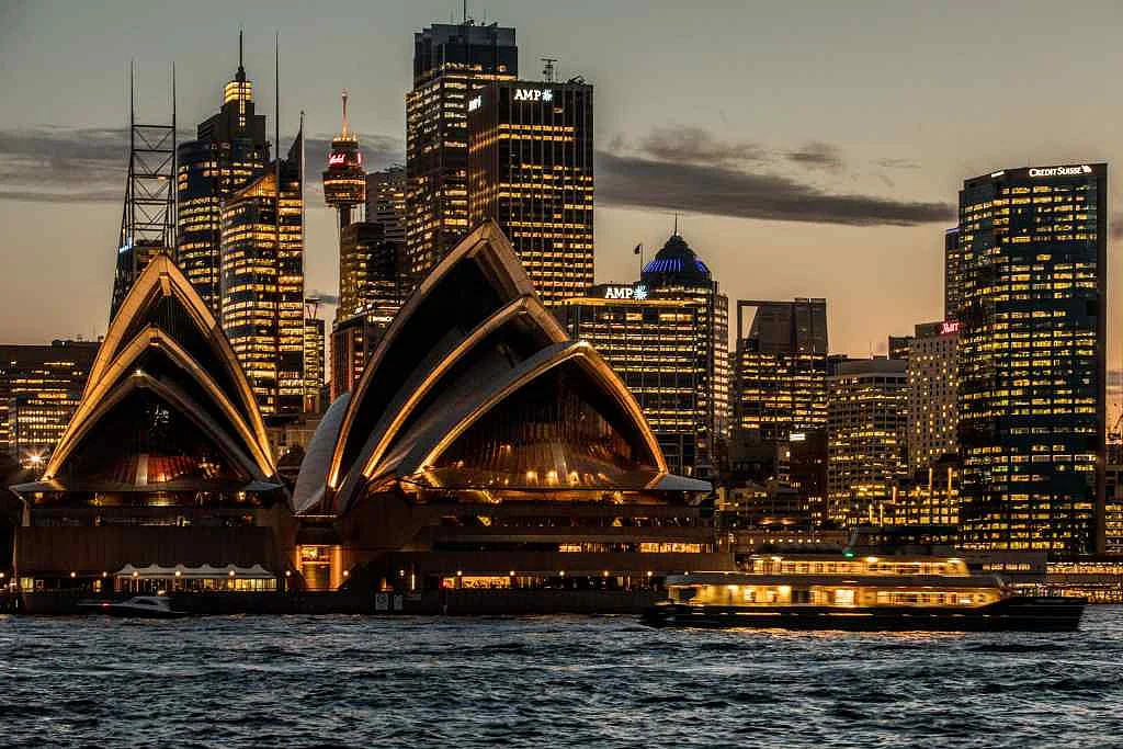 The Sydney Opera House, foreground, and buildings in the financial district stand illuminated at dusk in Sydney, Australia.