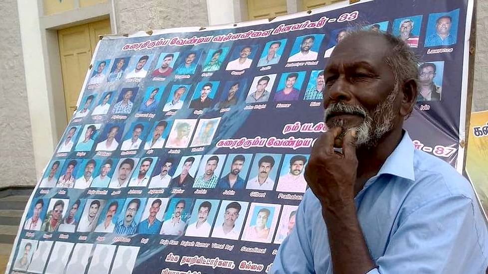 Fisherman Amaladasan stands beside a poster showing photos of 70 fishermen missing from Vallavilai village in Kanyakumari district since Cyclone Ockhi.&nbsp;