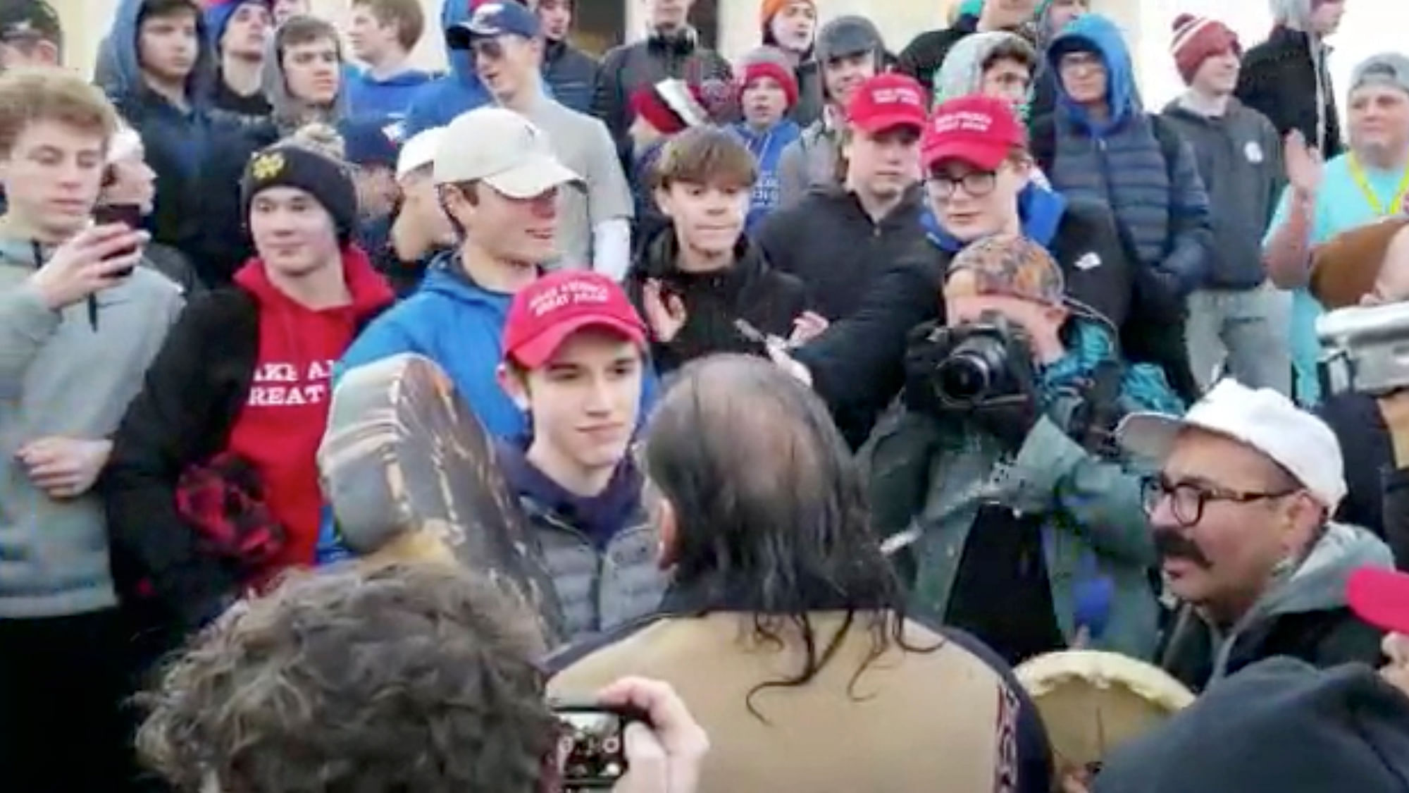 In this Friday, 18 January, image made from video provided by the Survival Media Agency, a teenager wearing a “Make America Great Again” hat, center left, stands in front of an elderly Native American singing and playing a drum in Washington.&nbsp;