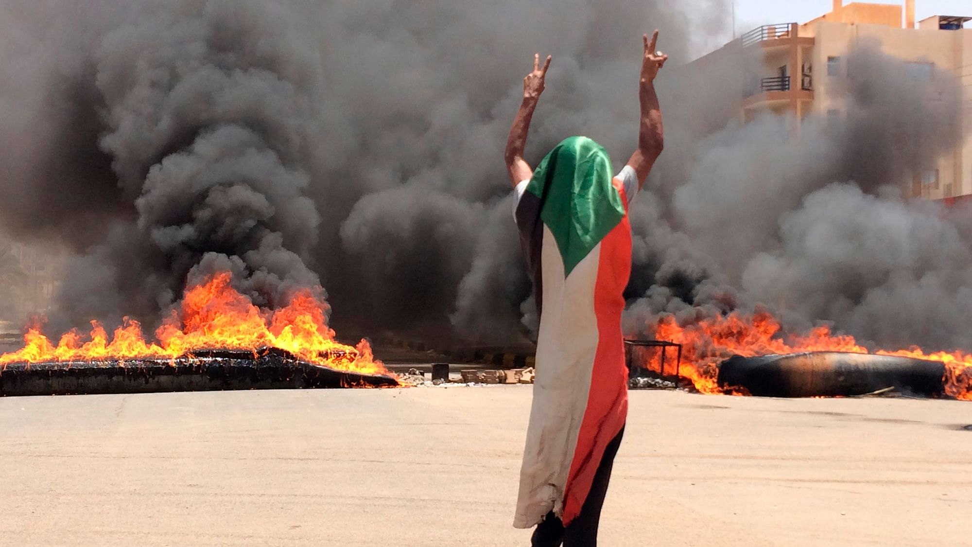 A protester wearing a Sudanese flag flashes the victory sign in front of burning tires and debris on road 60, near Khartoum’s army headquarters, in Khartoum, Sudan.