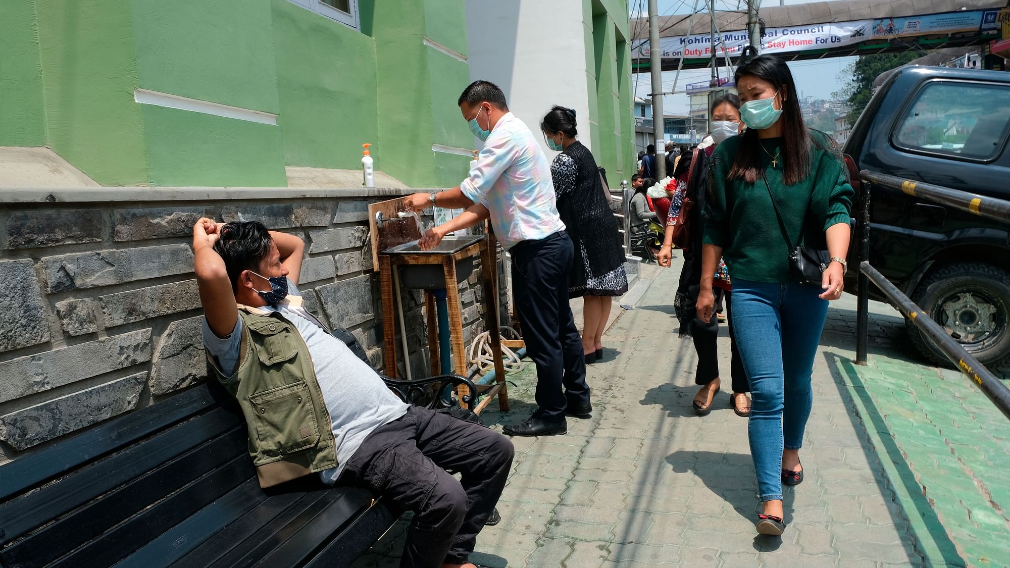 A man rests on a bench as pedestrian wash their hands at hand basins set up by the entrance of Oking Hospital in Kohima, capital of the northeastern Indian state of Nagaland, Monday, April 13, 202