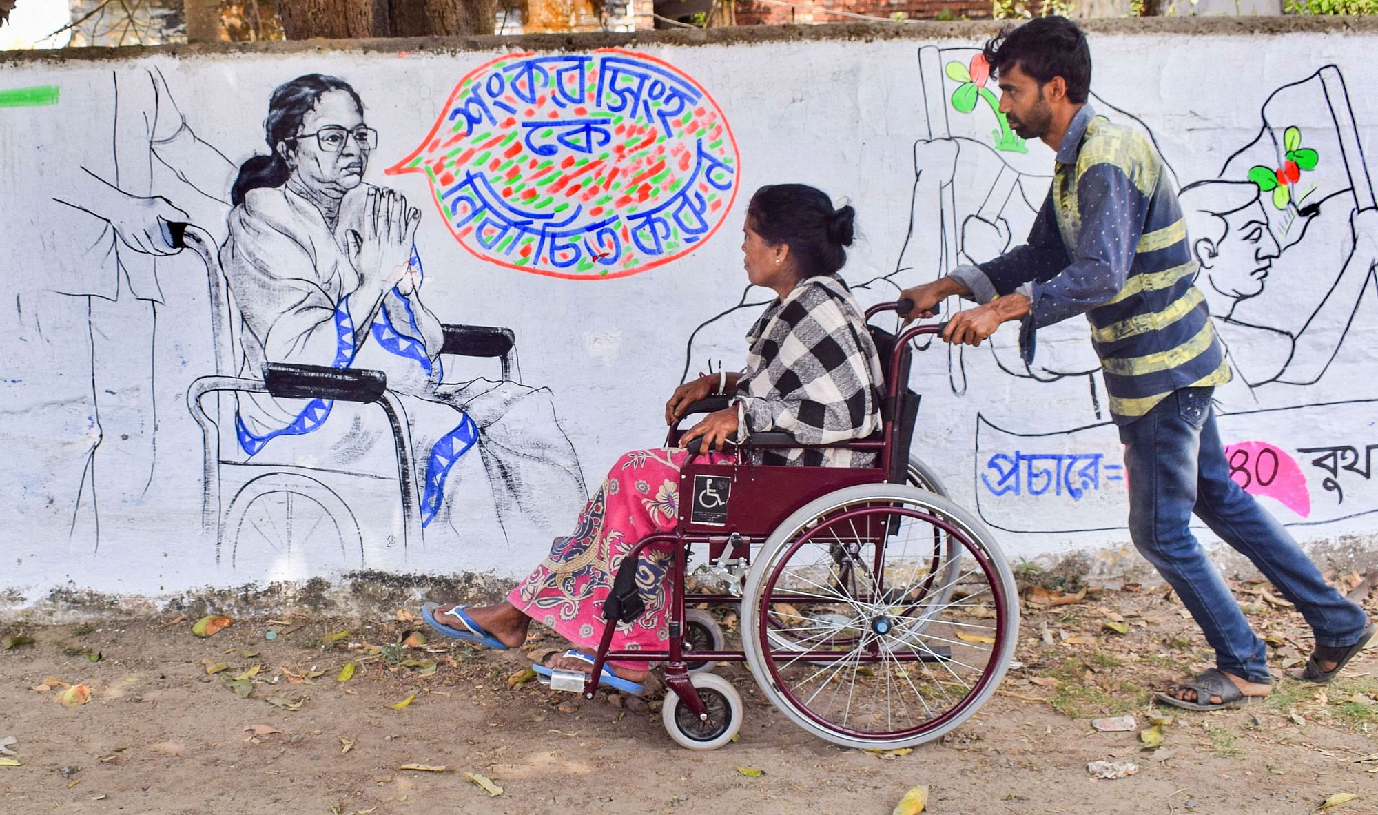 Nadia: A man pushes an elderly woman on a wheel-chair past a wall mural of West Bengal CM Mamata Banerjee, ahead of the upcoming state Assembly polls, in Nadia district, on Wednesday, 17 March.